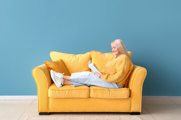 Mature woman reading book while sitting on sofa near blue wall