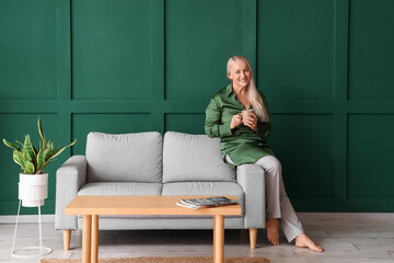 Obraz premium Mature woman drinking tea while sitting on sofa near green wall