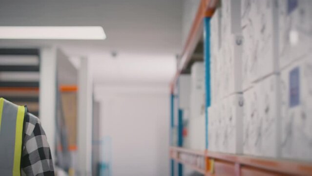 Camera tracks past warehouse shelves to show male worker wearing high vis safety vest in distribution warehouse smiling into camera - shot in slow motion