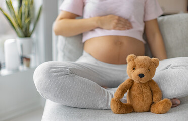 Pregnancy. Pregnant woman wearing maternity clothes relaxing on home sofa with teddy bear baby toy in focus for baby shower gift concept or expecting a child