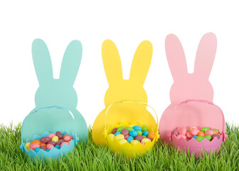 Three colorful wooden Easter Bunny Silhouettes in blue, yellow and pink with matching colored tiny easter baskets full of candy jelly beans sitting on green grass, isolated on white background.