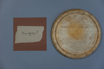 A round, empty wooden cutting board against a gray background. A piece of paper with the question Hungry against the brown square? Top view