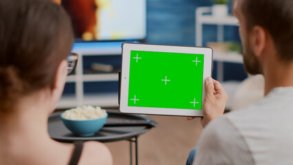 Closeup of couple holding digital tablet with green screen watching webinar and talking sitting on couch. Man and woman looking at touchscreen device with chroma key attending online course at home.