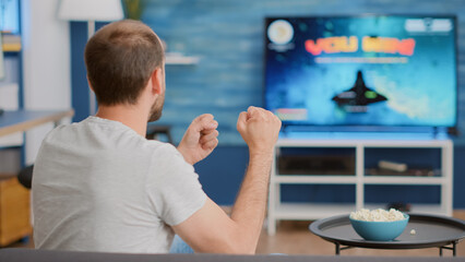 Closeup of man holding controller celebrating victory in online first person shooter game in front of big screen tv. Gamer doing celebration hand gesture happy with performance in gaming simulation.