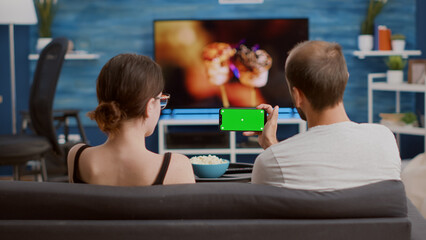 Static tripod shot of man holding smartphone with green screen watching online video content with girlfriend sitting on sofa. Couple enjoying social media videos on mobile phone with chroma key .