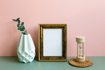 Blank picture frame, vase of plant, sandglass on green table. pink wall background. home interior