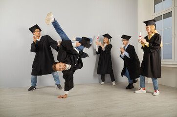 Congrats, grad. Group of excited university graduates in hats and robes dancing and having crazy fun on graduation day. Happy male student doing a break dance handstand and his friends are applauding