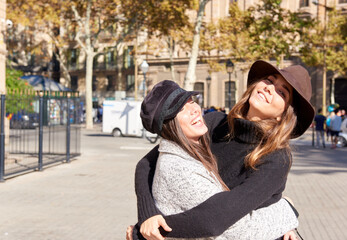 Female tourist friends wearing warm clothes hugging and laughing on city square on sunny day,...