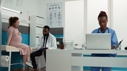 Medical assistant working on laptop in cabinet at appointment, helping male obstetrician with...