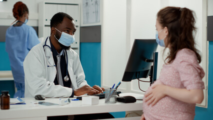 Fototapeta premium General practitioner examining woman with pregnancy belly during coronavirus pandemic. Male specialist talking to patient expecting child, giving medical advice and helping with maternity.