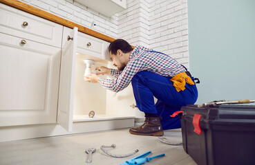 Service industry. Serious professionalmale plumber installs siphon under sink in kitchen of apartment. Side view of man in work uniform squatting near toolbox and repairing asink drain.