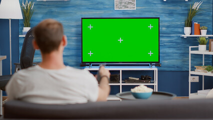 Man sitting on sofa looking at green screen on tv and switching channels in modern living room. Back view of person relaxing on couch using television remote zapping programs on chroma key display.
