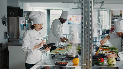 Authentic chef looking at food recipe on digital tablet screen, using online internet website to make gastronomy dish. Woman cook with device making professional cuisine restaurant meal.