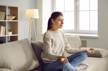 Calm young woman sit on sofa in lotus position with mudra hands meditate practice yoga. Relaxed...