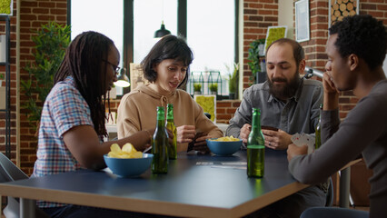 Diverse group of friends playing cards at fun gathering, enjoying competitive board games for entertainment. Men and women drinking beer and eating snacks to play stratgey game.