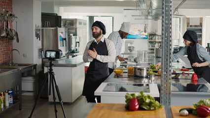 Professional cook filming cooking show video on camera, working in restaurant kitchen. Authentic male chef recording culinary TV program with professional gastronomy, making gourmet dish.
