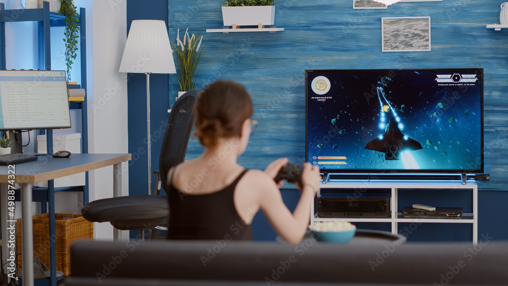 Wall mural Gamer girl holding wireless controller playing console video game first person shooter on tv while sitting on sofa in modern living room. Young woman enjoying online action game while eating popcorn.