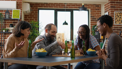 Multiethnic group of people having fun with chess game, playing match on chessboard. Cheerful friends meeting in apartment to enjoy board games play and alcoholic drinks together.