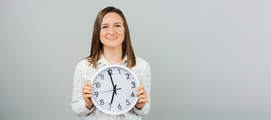 Smiling at the camera young woman is holding a white round wall clock. Panorama studio shot over...