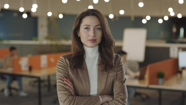 Beautiful caucasian businesswoman in a good mood looking into camera and smiling indoors. Portrait of handsome female manager standing with crossed arms in the office room