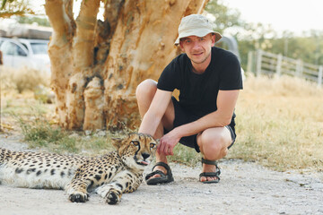 Male tourist in casual clothes and hat sitting and petting a cheetah outdoors