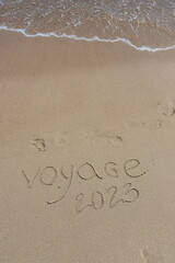The inscription voyaje 2022 in French, travel 2022, on the sand by the water and the rising wave, seashore beach vacation by the sea
