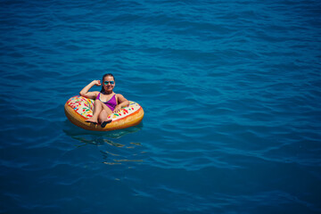 A young beautiful girl in a bright swimsuit lies on a large inflatable ring and floats on the blue sea on a bright sunny summer day