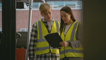 Female team leader with clipboard in warehouse training male intern standing by fork lift truck - shot in slow motion - Powered by Adobe