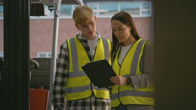 Female team leader with clipboard in warehouse training male intern standing by fork lift truck - shot in slow motion