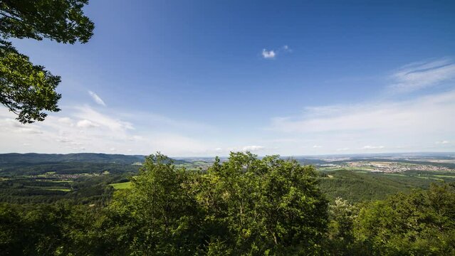 Timelapse image of clouds in landscape. The clouds pass over the camera. In the foreground are green deciduous trees. In the valley are various residential areas and villages. The sky is strong blue.