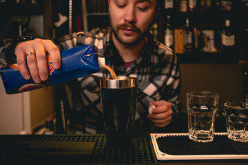 Bearded bartender pouring orange juice from carton to shaker, preparing cocktail
