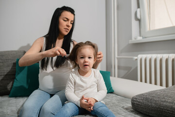 Fototapeta premium Mother and daughter sit on the sofa and have a fun time. A pregnant mother does hairdressing for her little daughter and combs her with a hairbrush. The kid looks at the camera and sits patiently.
