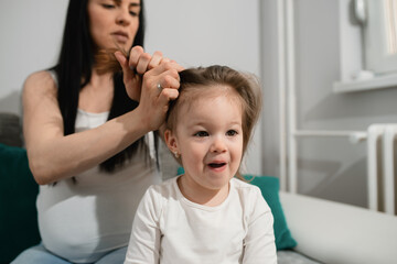 Caucasian pregnant woman makes hairdo for her little daughter. Blacak-haired female combes the child's hair with her hands while the little girl sits on the couch. 
