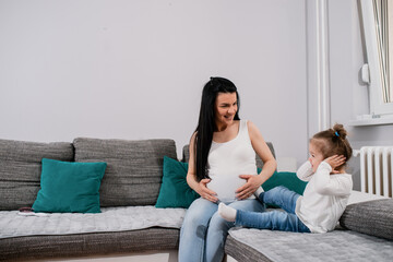 Good-looking black-haired pregnant caucasian female touches big tummy with both her hands,smiles and looks at her daughter who covers her ears with hands.Cute little girl enjoys spending time with mom