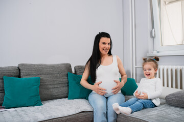 Beautiful pregnant mom and her wonderful girl joy spending time together. They are laughing on the couch in the living room. A woman touches her tummy with both hands while little girl giggles. 