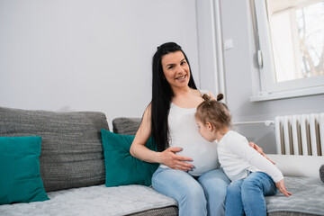 A little girl with ponytails kisses her mother's tummy while they sit on the gray sofa in the living room. A pregnant black-haired white woman smiles and touches her belly with her right hand. 