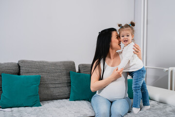 Cheerful mother and daughter are on the sofa. The pregnant mom's face turned to her daughter's face and a little girl with ponytails looks at the camera and smiles.A cute child stands on the couch nex