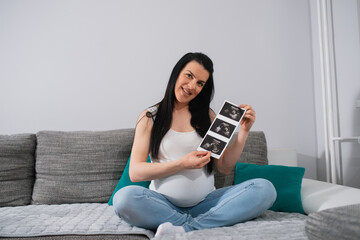 Well-looking pregnant female proudly shows ultrasound pictures of her unborn baby. A smiling caucasian woman sits on a gray sofa and looks right at the camera. Green pillows are behind her back. 