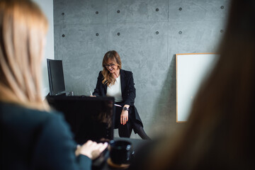 A smiling, attractive middle-aged female trainer with eyeglasses takes notes in class in a modern office. Next to her is a whiteboard for presentation. Two unrecognizable girls listen to lectures. 