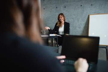 A smiling middle-aged Caucasian female instructor has a conversation with seminar participants in a corporate office. Whiteboard is beside her and a gray wall behind her. She holds pen in right hand. 