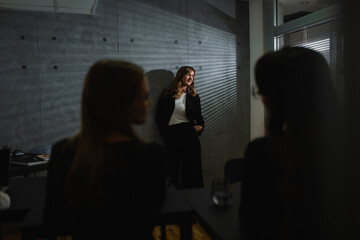 A senior, smiling female teacher leans against a gray wall in a darkened room.Two blurred caucasian female students are facing each other and talk while the teacher thoughtfully looks out the window. 