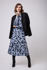 High fashion photo of a beautiful elegant young woman in a pretty long blue dress with floral pattern, black jacket, boots posing on white background. Slim figure, hairstyle, studio shot.