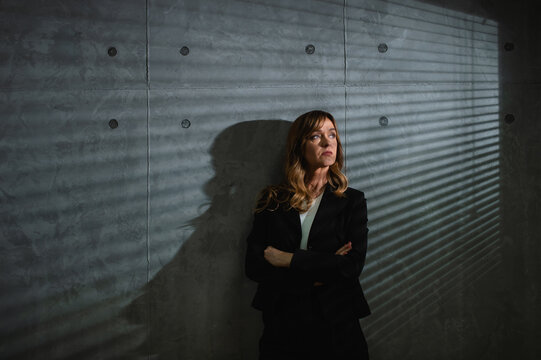 A dreamy senior brown-haired formal clothed caucasian woman stands in front of gray background in darkened room with crossed arms. Blins and her shadows are visible on the wall. 