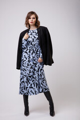 High fashion photo of a beautiful elegant young woman in a pretty long blue dress with floral pattern, black jacket, boots posing on white background. Slim figure, hairstyle, studio shot.