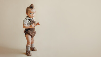 an angry kid with a funny grimace in a vintage jumpsuit stands on a beige background and holds a cookie in his hand