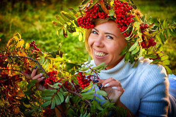 Cute girl in a white dress, wreath of rowan branches and whith a bouquet of red berries in nature in a clearing in the forest. Photo shoot on green grass in sunny autumn day