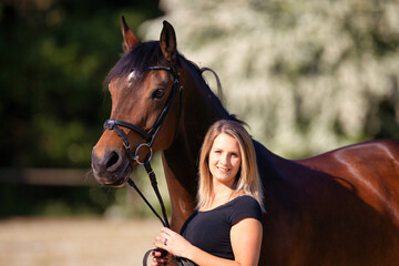 Obraz premium Young woman stands next to her horse and looks into the camera, horse looks to the left with ears pricked..