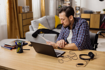 A bearded man works at home at a desk on which lie a phone, headphones, laptop, tablet and calendar. The busy brunet carefully reads the documents held in his hand about a new business partner.