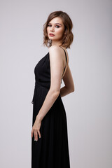 Fototapeta premium High fashion photo of a beautiful elegant young woman in a pretty black evening party dress with a deep neckline posing on white background. Slim figure, hairstyle, studio shot. 