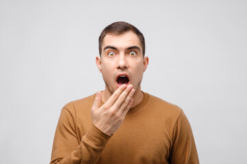 portrait of surprised caucasian brunette man with hand covering his open mouth in surprise and big dumbfounded eyes on grey background